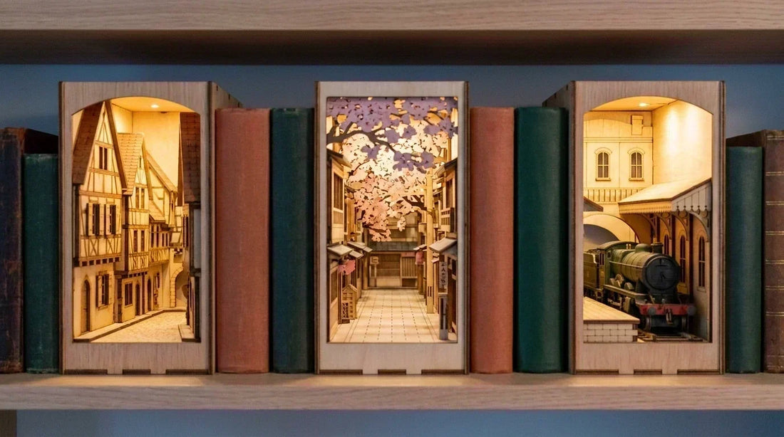 Trois book nooks voyage alignés sur une étagère de bibliothèque : ruelle alsacienne, ruelle japonaise et quai de gare londonien, éclairés par leurs LED.